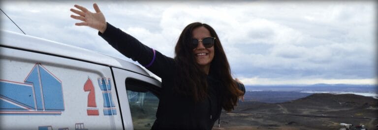 woman smiling and waving in front of a rent.is camper van