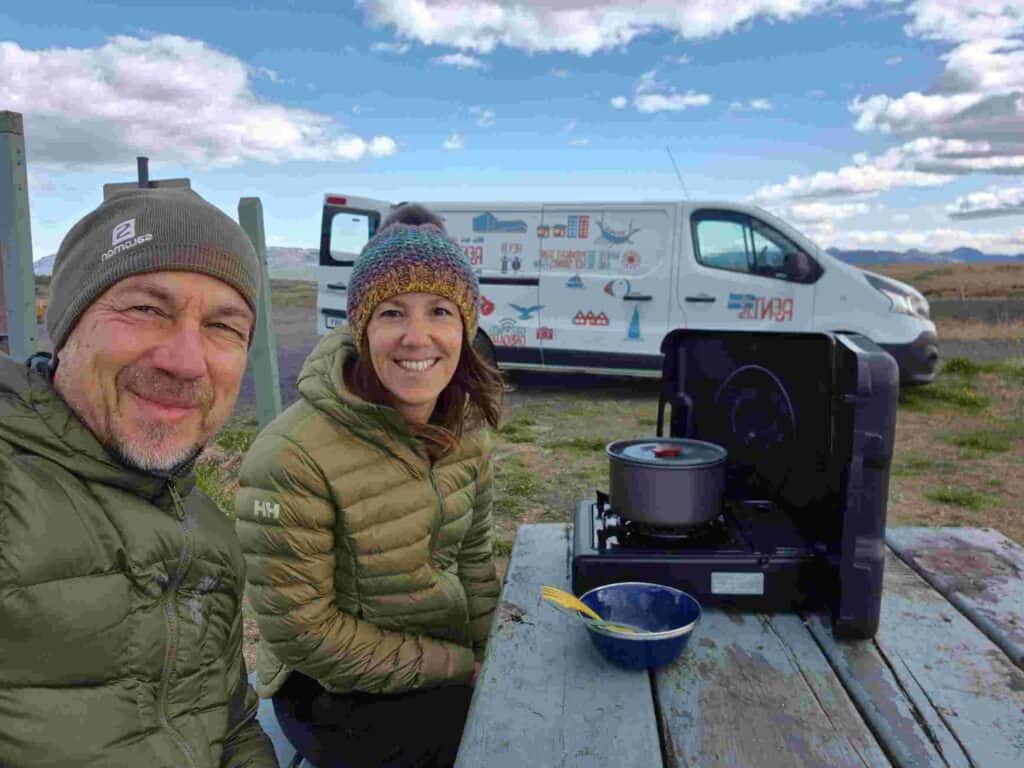A couple cooking food on a picnic table next to renault trafic