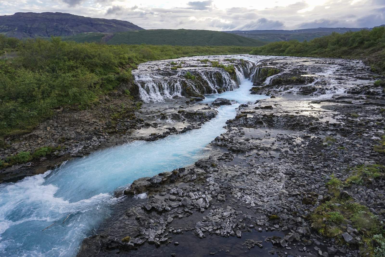 Brúarfoss waterfall pouring through a narrow gap into bright blue water, seen from the bridge