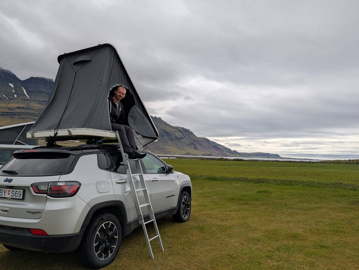 Man sitting in the open rooftop tent on a silver Jeep Compass parked on grass by a fjord in Iceland