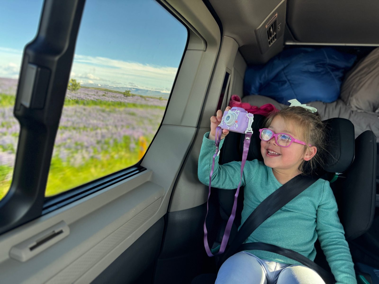Smiling girl with a toy camera in her car seat in the camper van, with lupin fields outside the window